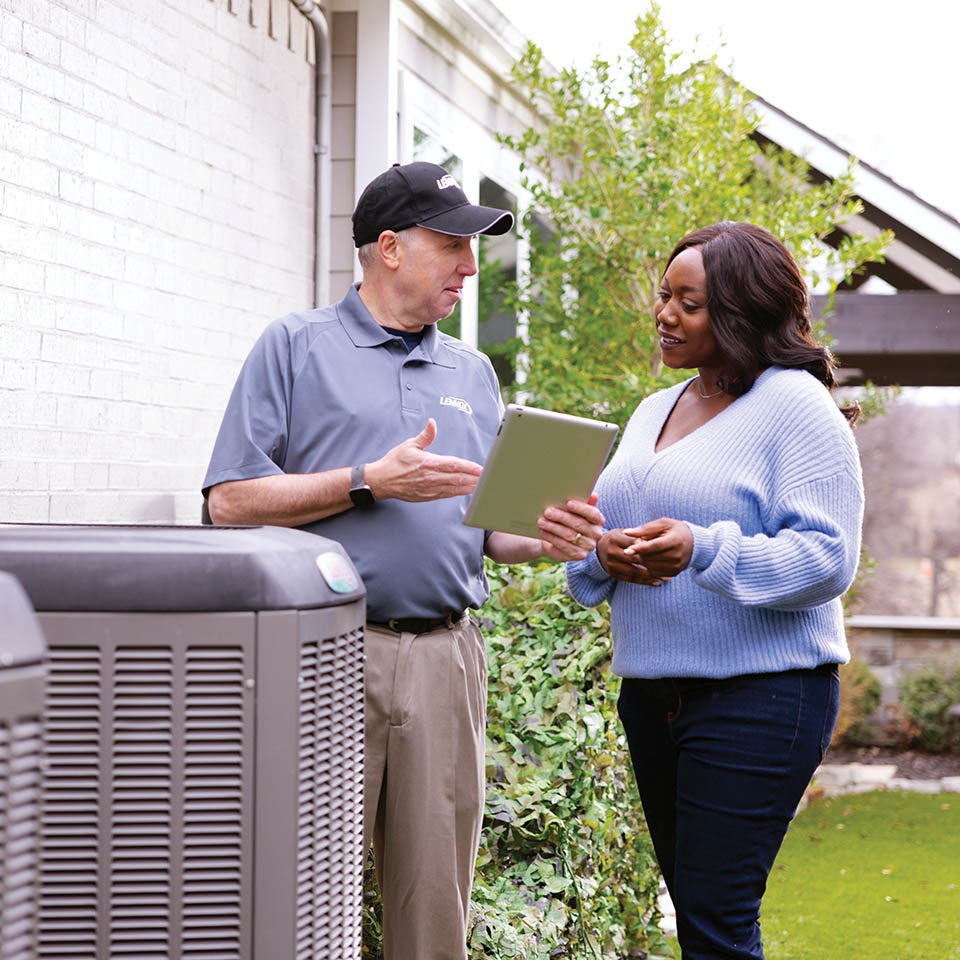 HVAC technician and homeowner standing next to an outdoor air conditioning unit, while the tech uses a tablet to explain information to the woman.