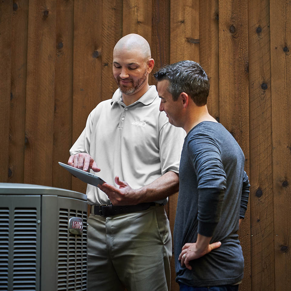 HVAC technician and homeowner standing next to a heat pump, while the tech uses a tablet to explain information to the man.